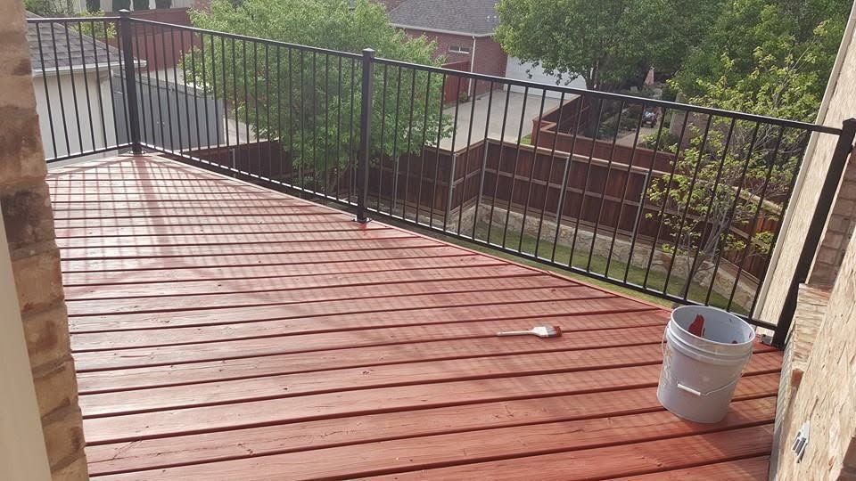 Wooden deck with black railing and a gray bucket on the right, overlooking a leafy yard.