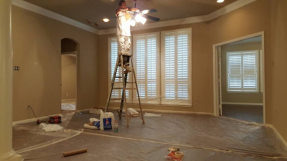 Living room under renovation with plastic-covered floor, ladder, and worker painting a vaulted ceiling.