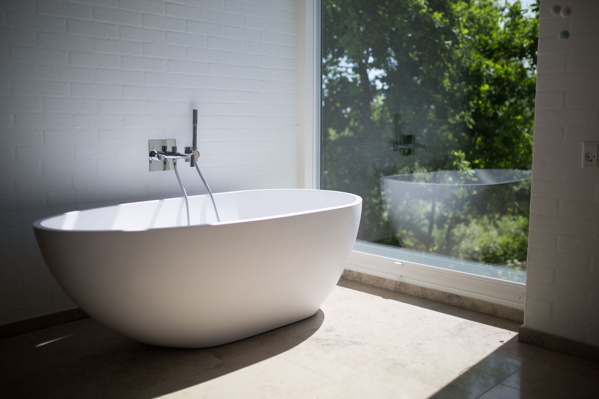 Modern bathroom with a white freestanding bathtub beside a large window and wall-mounted faucet