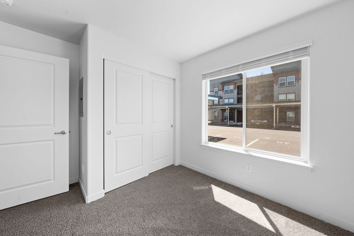 Empty white bedroom with carpet, two closet doors, and a window letting in bright sunlight.