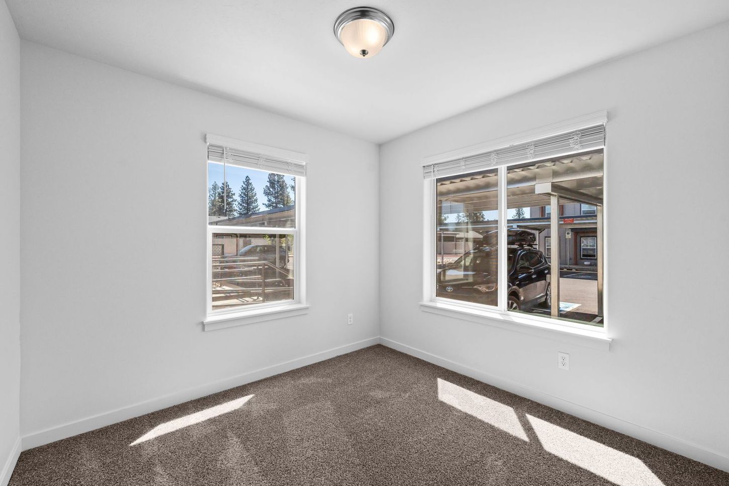 Empty white bedroom with two windows and gray carpet, sunlight streaming in.