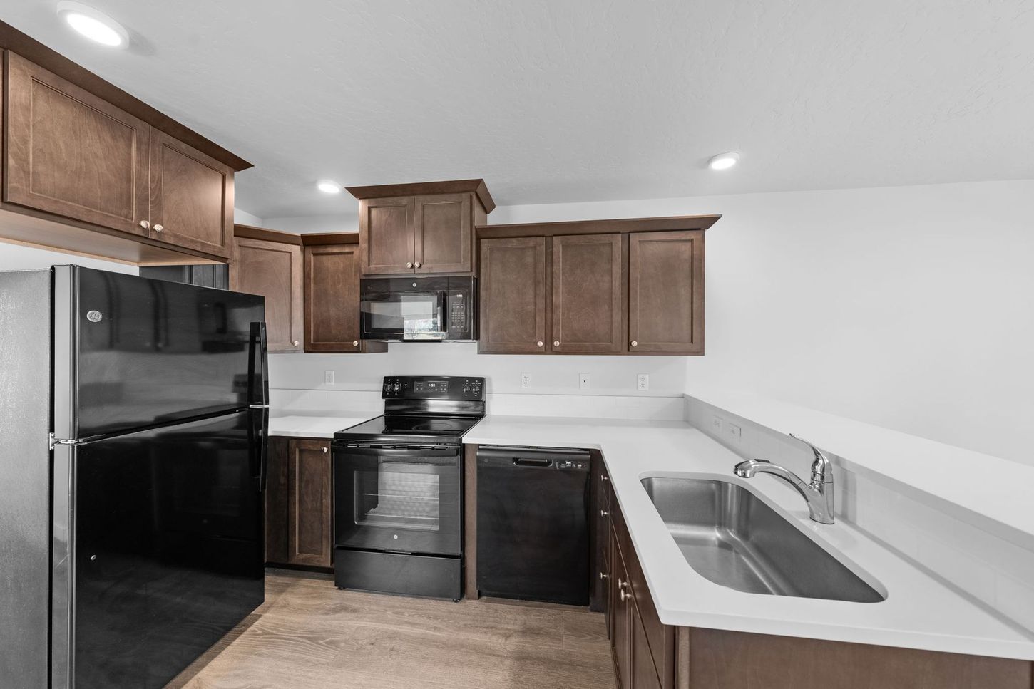 Compact kitchen with dark wood cabinets, black appliances, and white counters with a sink.