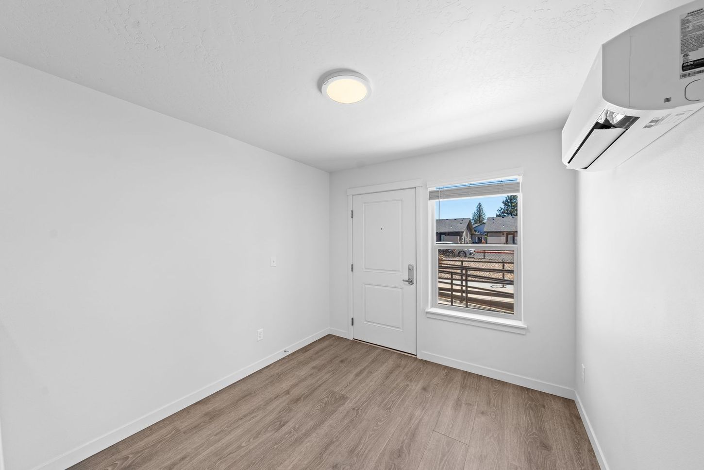 Empty white bedroom with wood flooring, a window, closet door, and wall-mounted air conditioner.