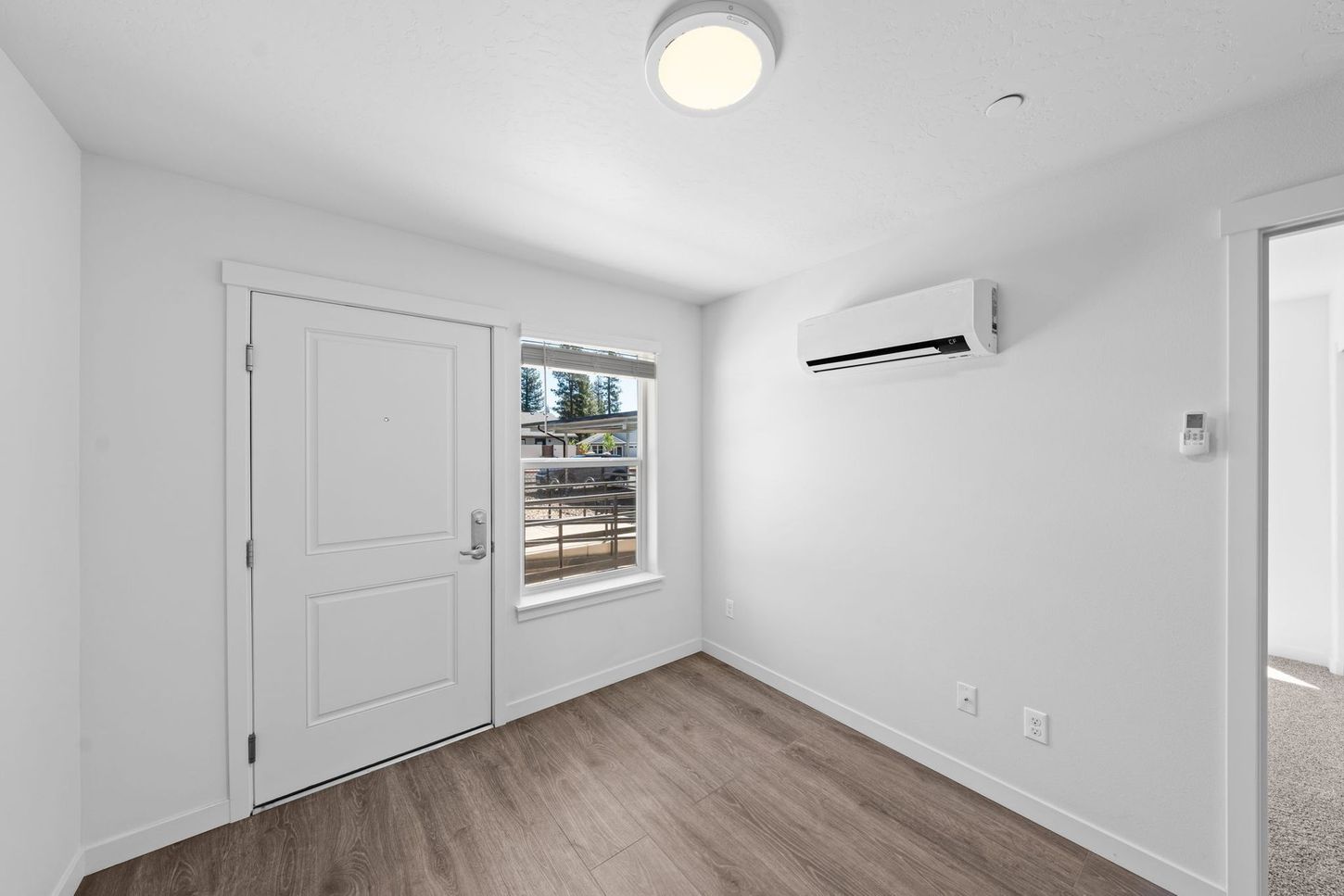 Small white room with gray carpet, a windowed door, wall AC unit, and ceiling light.