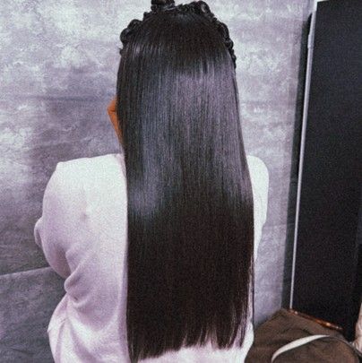 A person with long, straight, dark hair styled with a partial top knot, wearing a light-colored top against a gray wall.