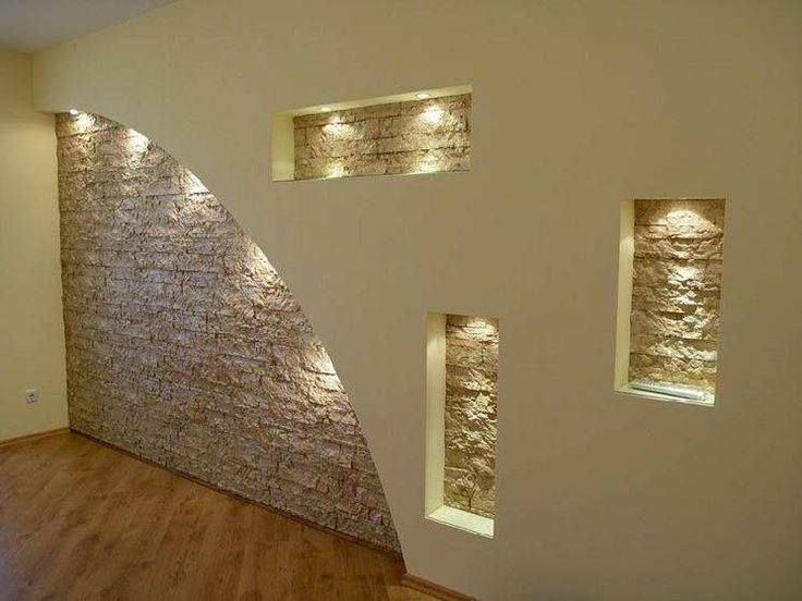 Parete beige con elementi in pietra e inserti, illuminata da faretti.