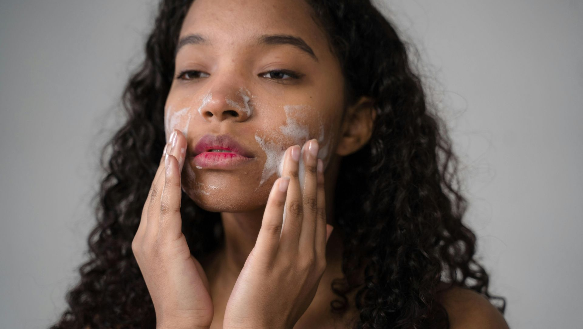Woman applying facial cleanser, hands on cheeks, eyes closed, white cream visible.
