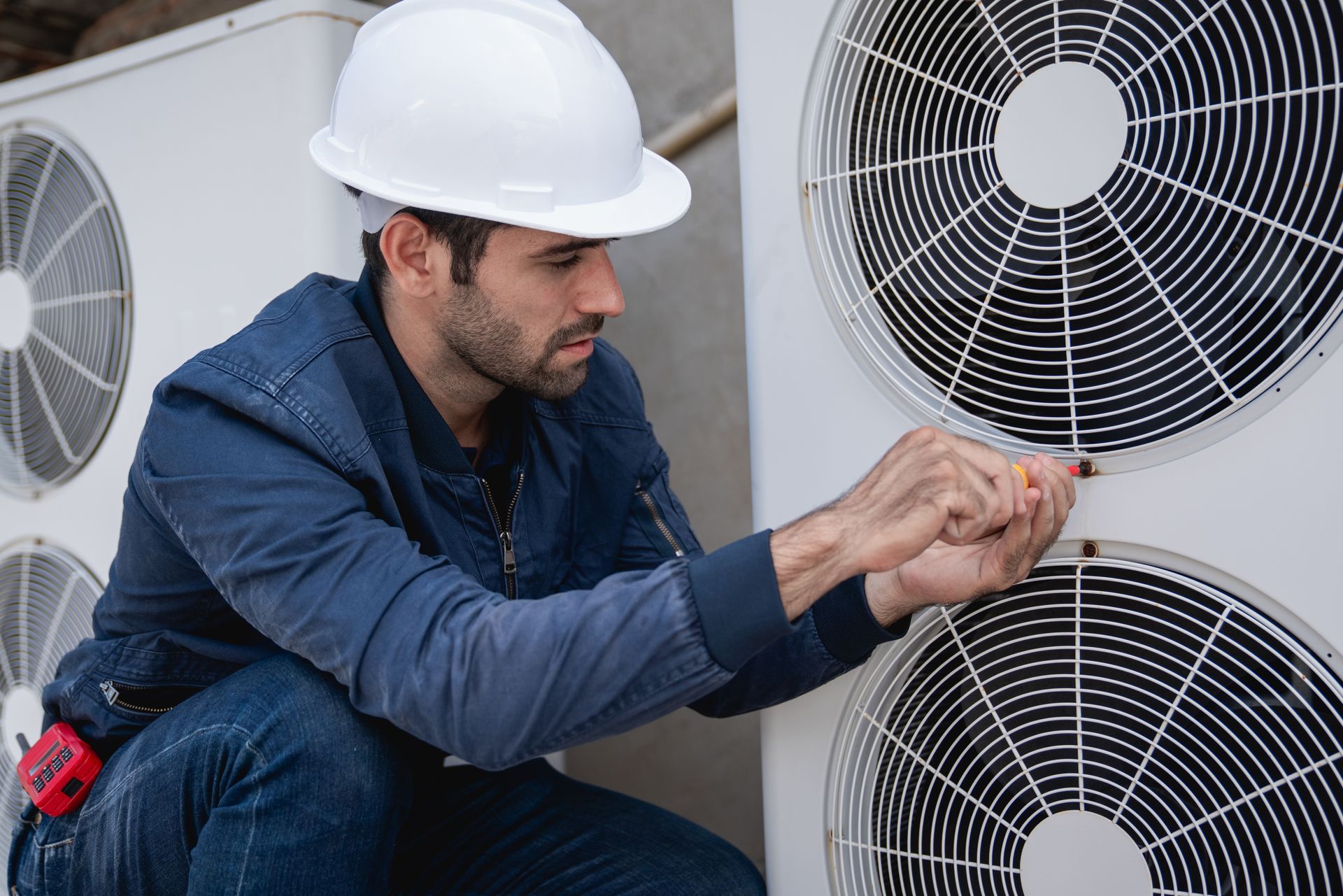 A person in a hard hat and blue work clothes using a tool to repair an outdoor air conditioning unit.