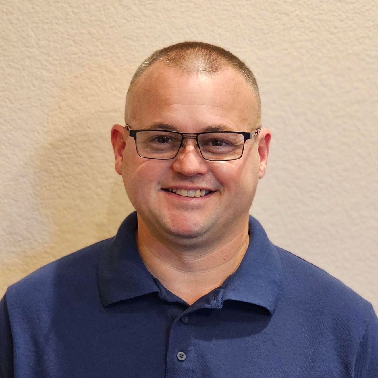 A person with short hair and glasses wearing a blue polo shirt, smiling against a neutral beige wall.