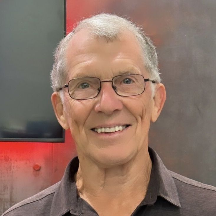 A person with short, gray hair and glasses smiles while wearing a collared shirt against a gray and red background.