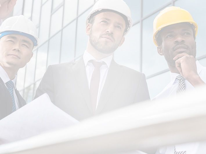 Three professionals wearing hard hats review blueprints in front of a modern glass building.