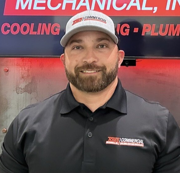 A smiling person in a black polo shirt and grey cap stands in front of a sign for a commercial mechanical company.