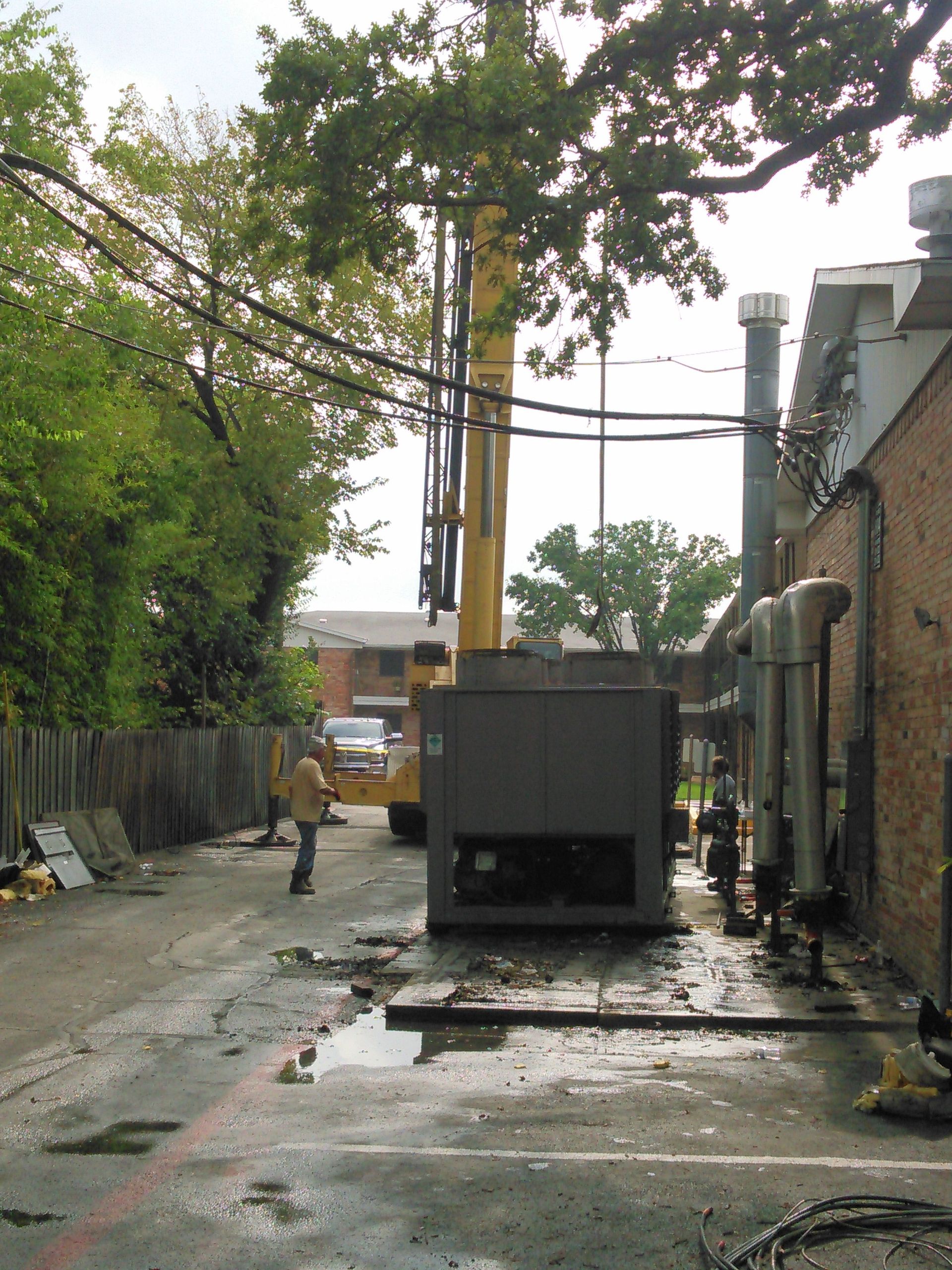 A crane lifts a large industrial HVAC unit in a narrow alleyway next to a brick building.