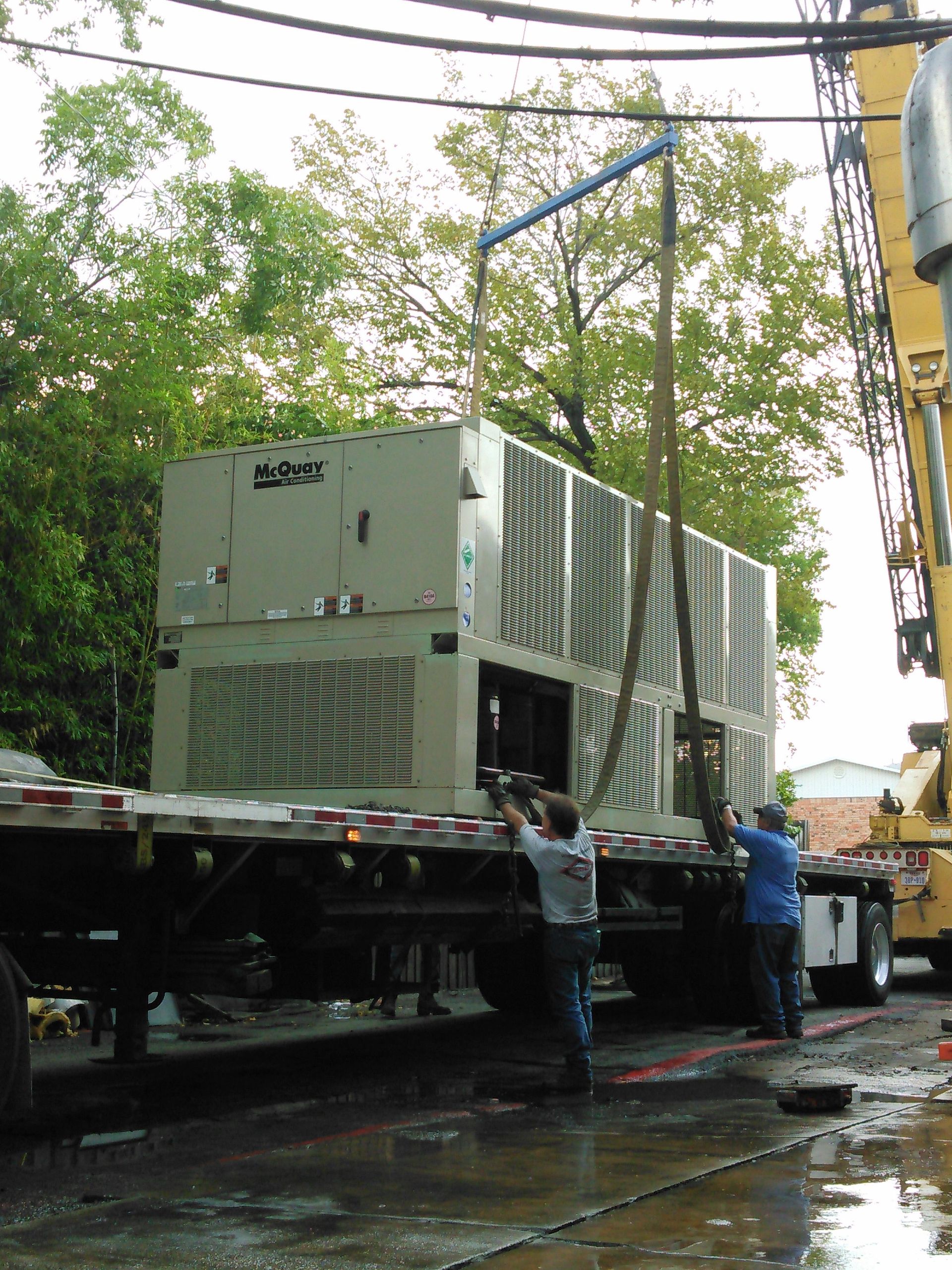 Two people guide a large, beige industrial HVAC unit being lifted off a flatbed truck by a crane.