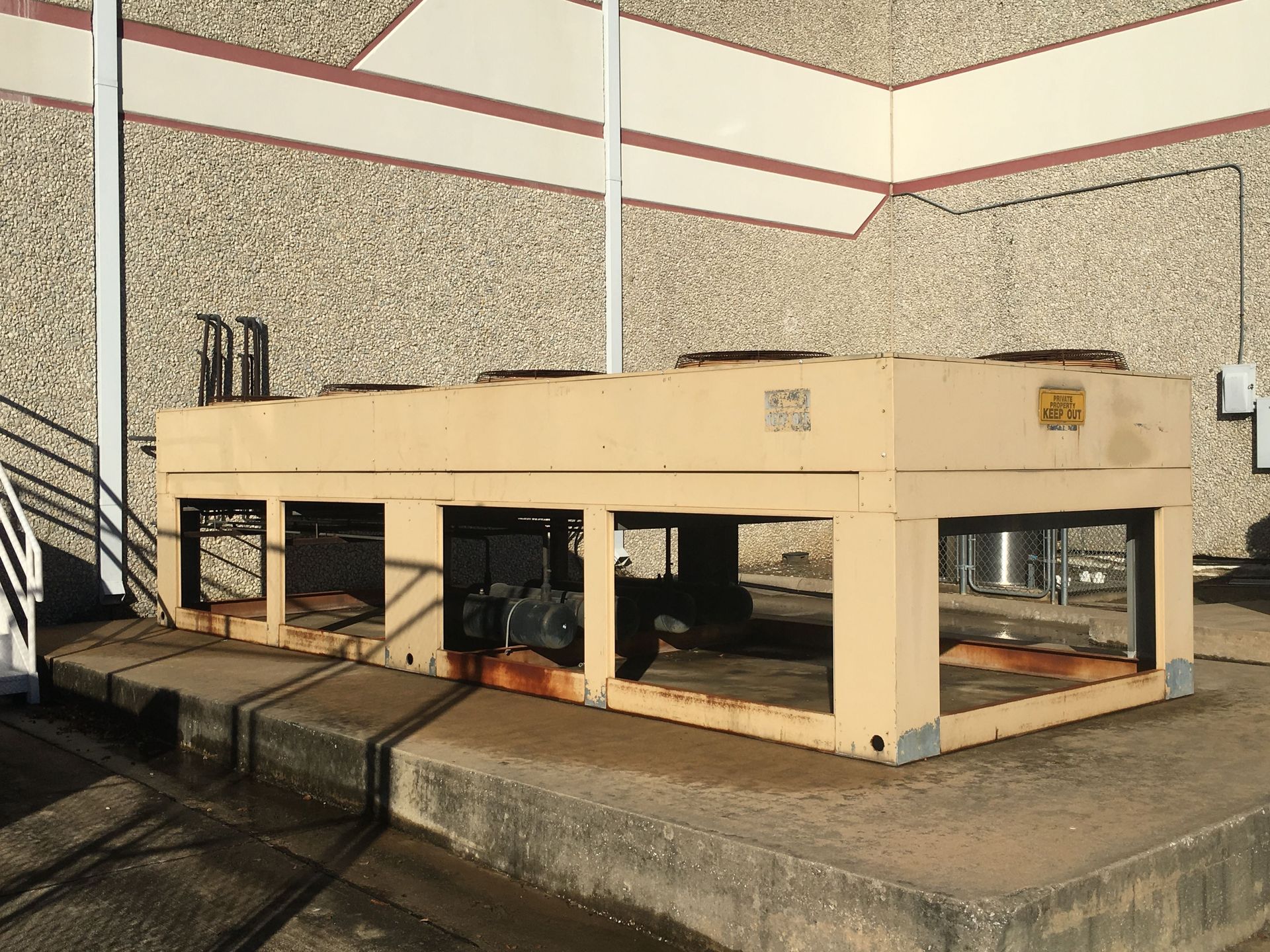A large, beige, rectangular outdoor industrial cooling unit sits on a concrete pad against a textured building wall.