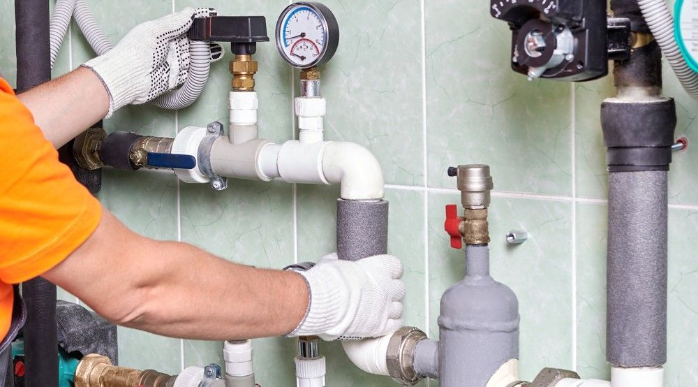 A person in an orange shirt and gloves works on white plumbing pipes mounted on a green tiled wall.