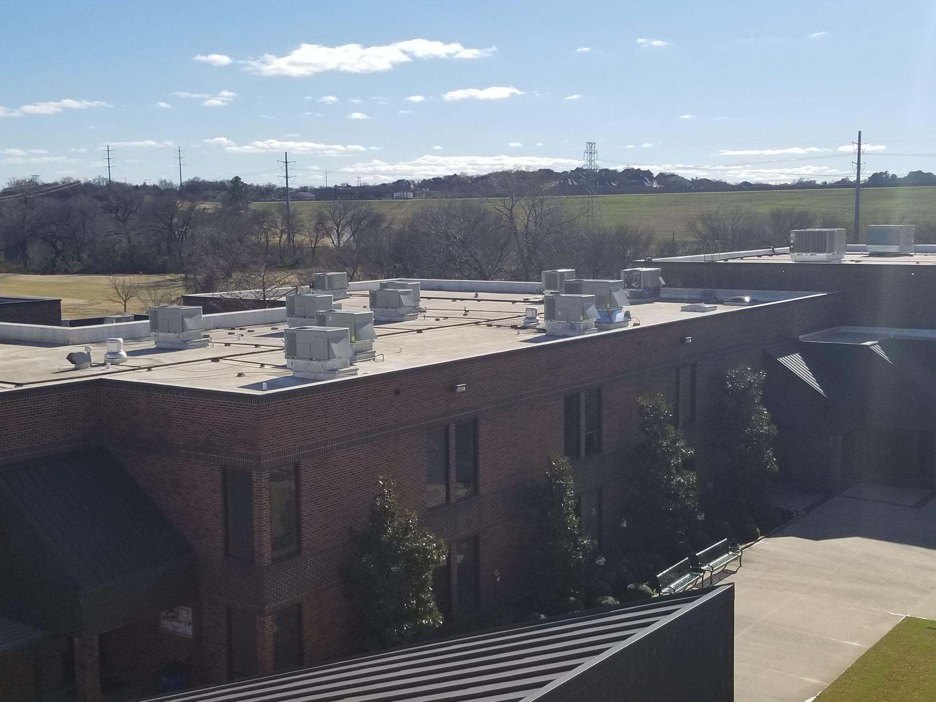 A multi-story red brick building with multiple HVAC units on its flat roof under a blue sky with scattered clouds.