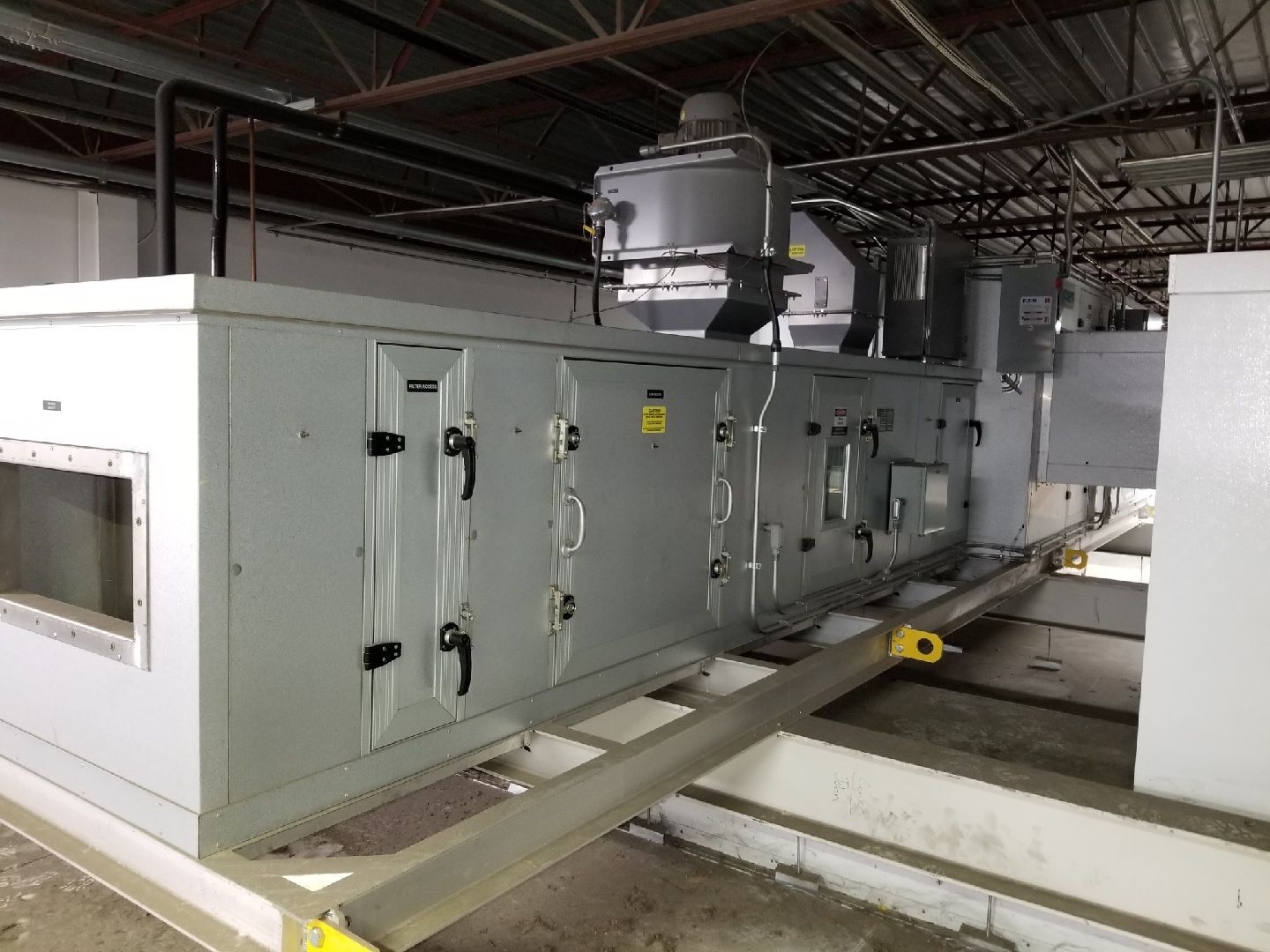 Large, light-gray industrial HVAC air handling unit mounted on metal beams in an open-ceiling mechanical room.