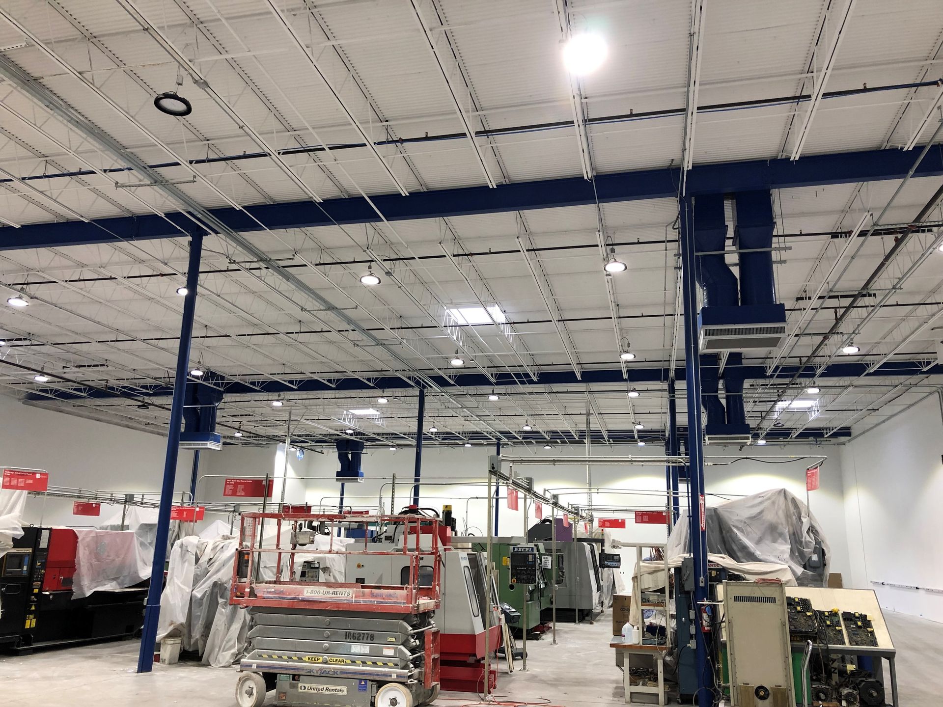 Large industrial warehouse with high ceilings, exposed beams, lighting, and heavy machinery on a concrete floor.