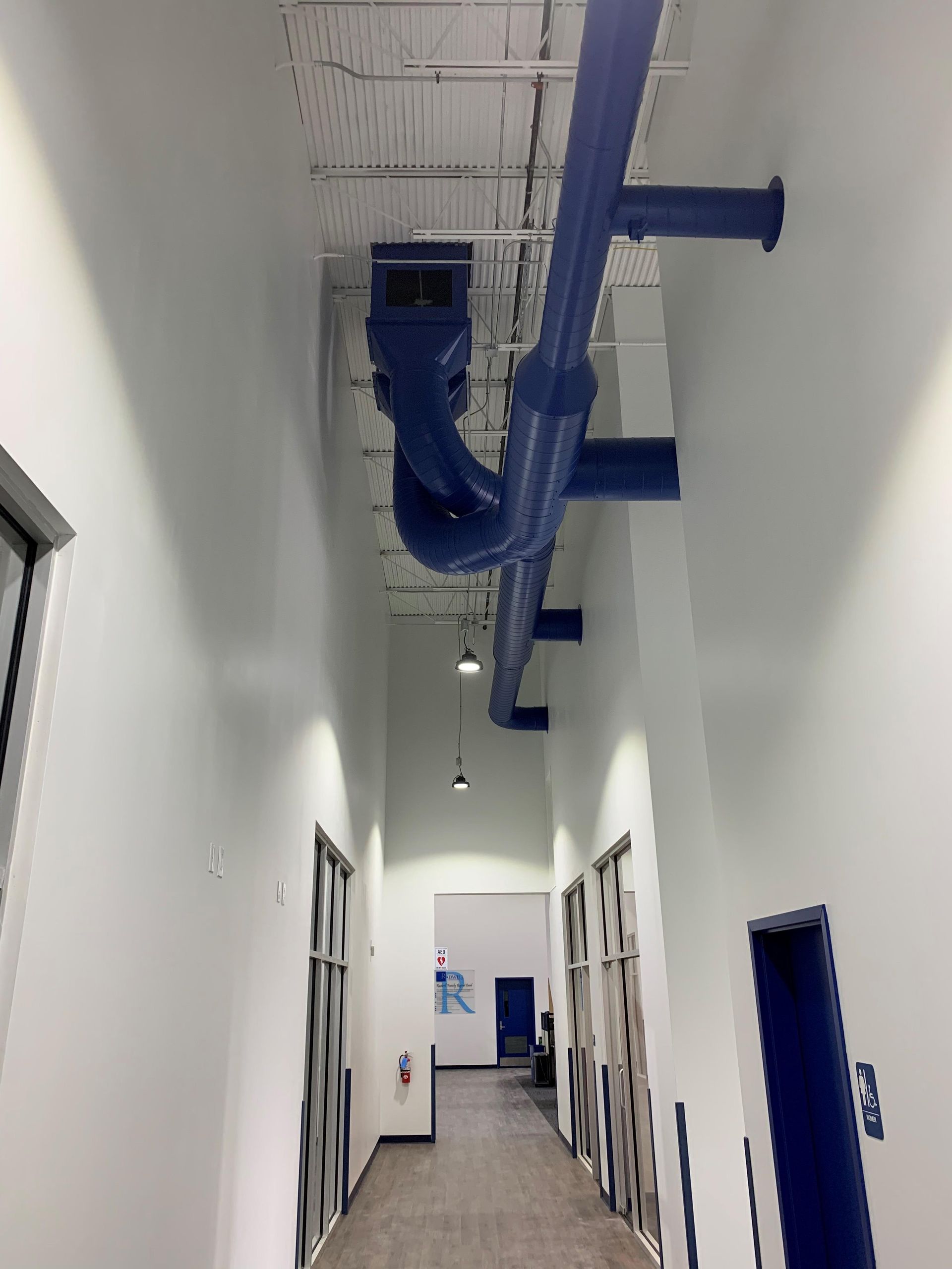 An indoor hallway with white walls and a high, exposed ceiling featuring large, blue-painted industrial pipes and ducts.