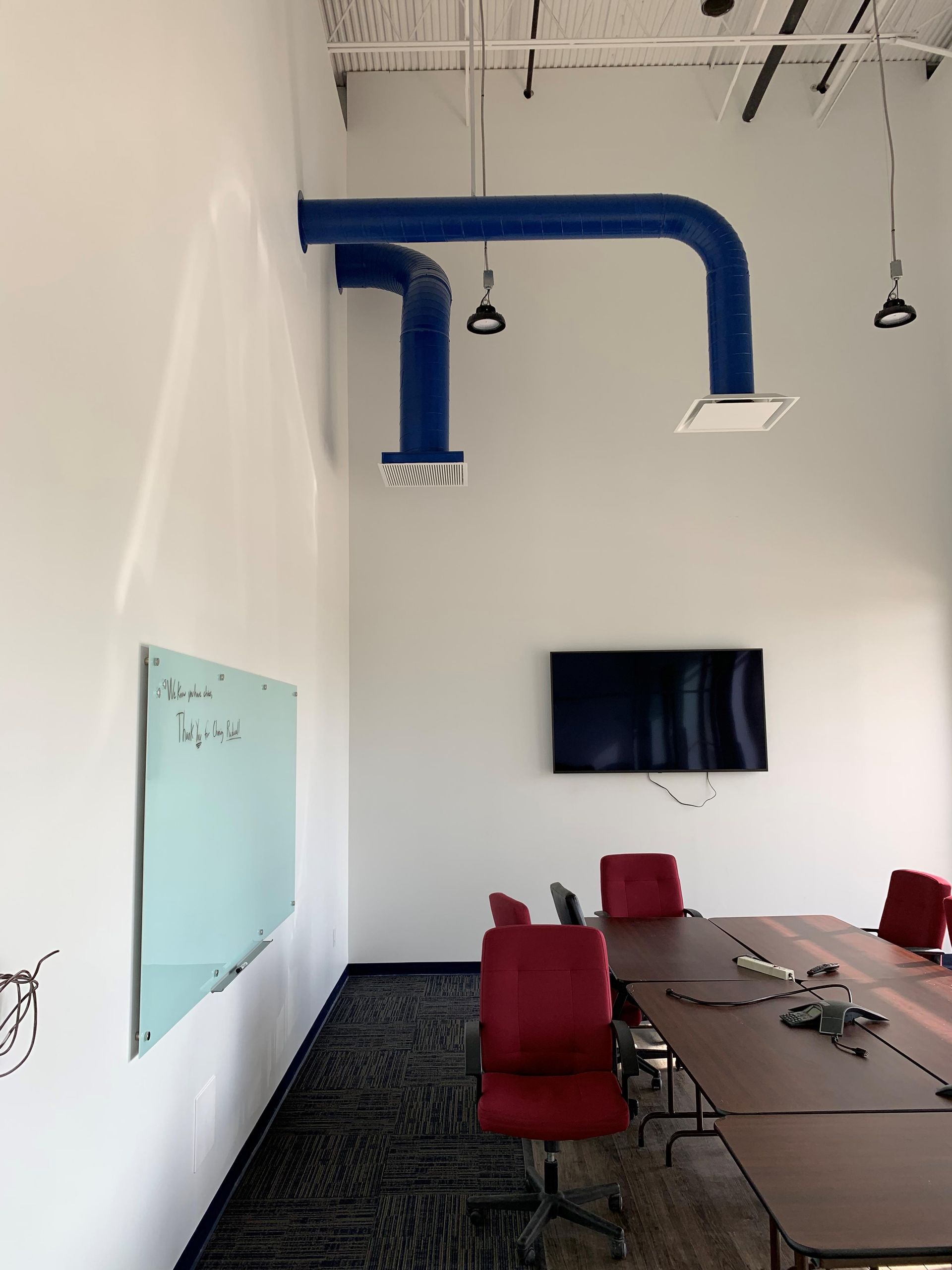 A conference room with a wooden table, red chairs, a wall-mounted TV, a glass whiteboard, and two blue ceiling vents.