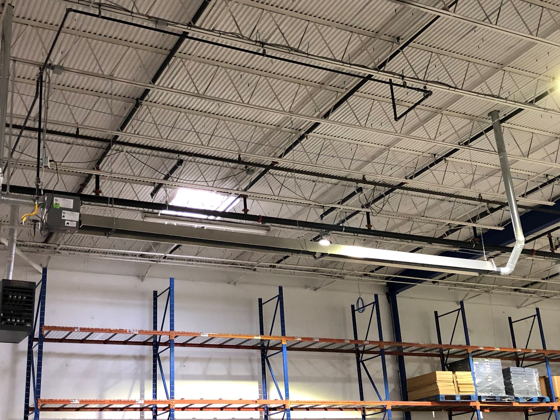 An overhead industrial infrared tube heater suspended from the ceiling above tall warehouse shelving.