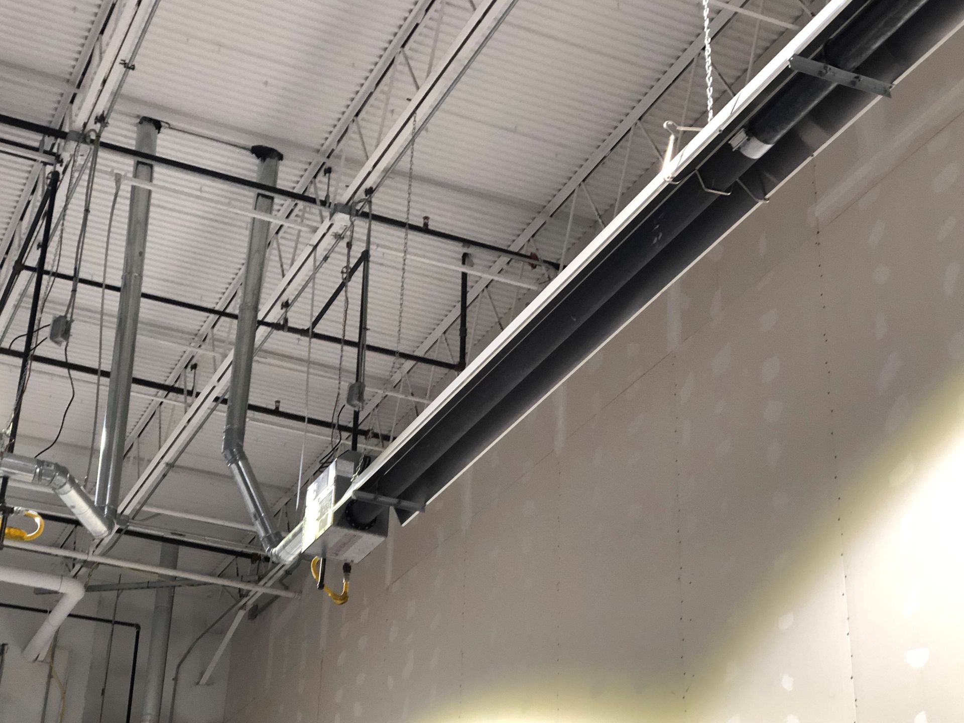A metal beam suspended from a warehouse ceiling, with chains, electrical conduits, and ductwork visible above.