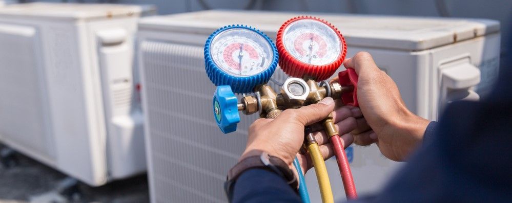 A technician holds a colorful HVAC manifold gauge set while servicing outdoor air conditioning units.