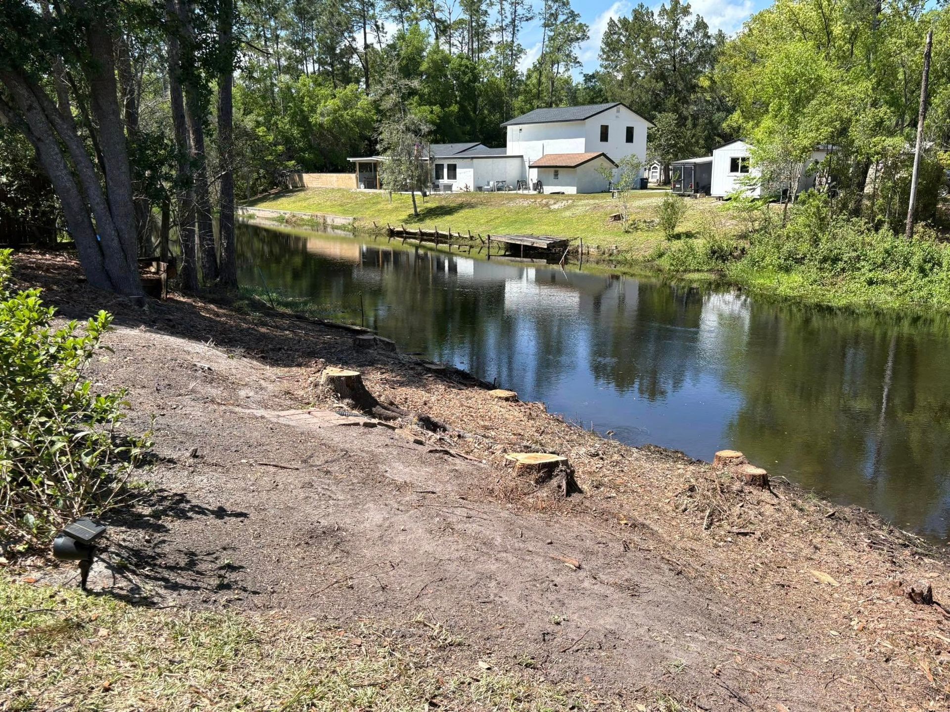 Wooded riverbank with calm water, trees, and a white house across the shore