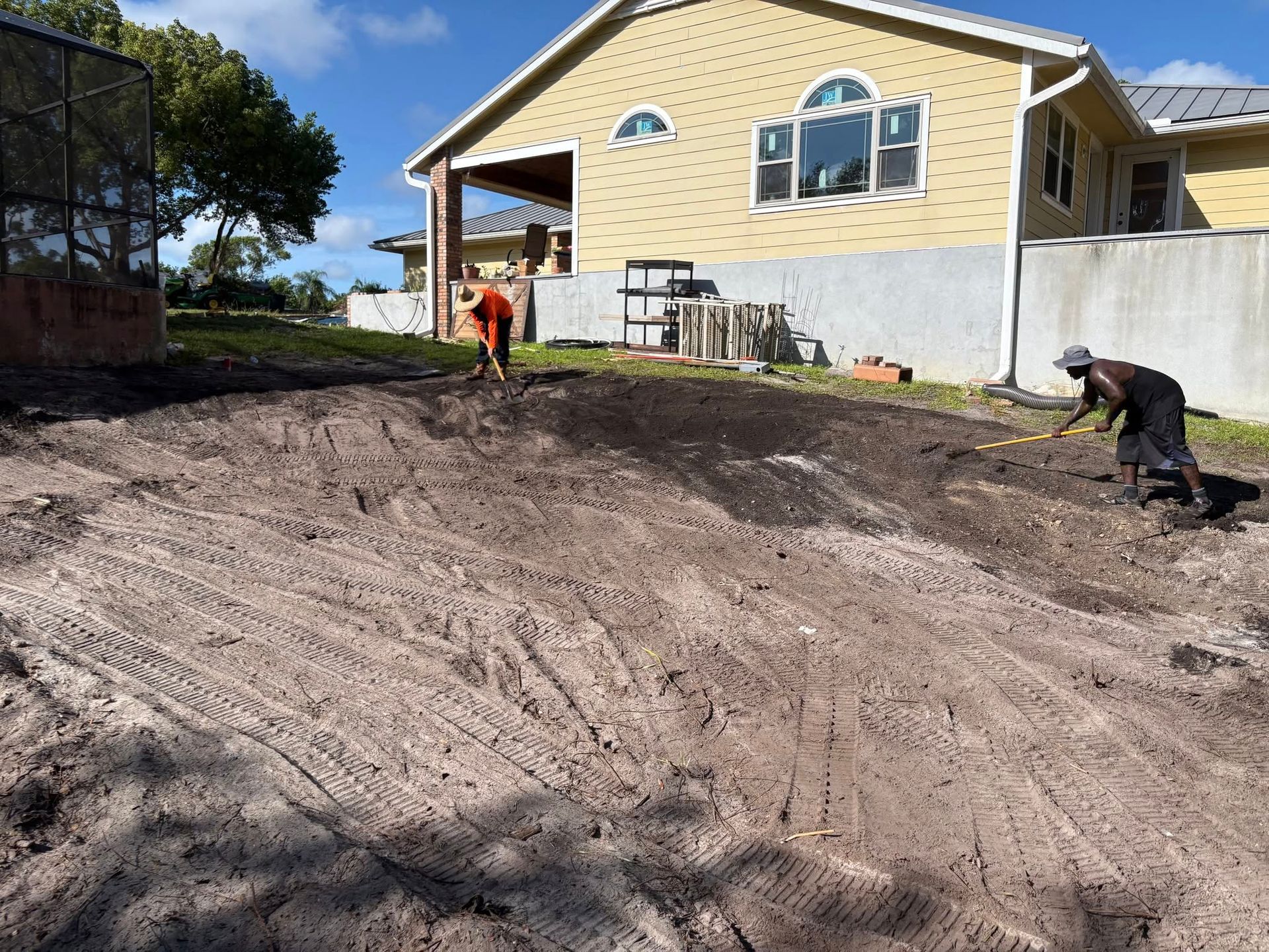 Two workers grade dirt in front of a yellow house on a sloped yard.