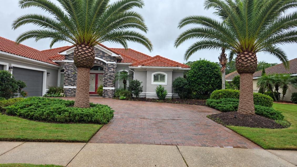 Mediterranean-style house with red tile roof, palm trees, and a brick driveway framed by lush landscaping