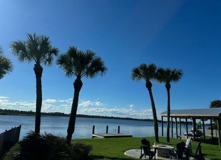 Palm trees beside a calm waterfront under a clear blue sky, with a grassy patio and chairs in the foreground