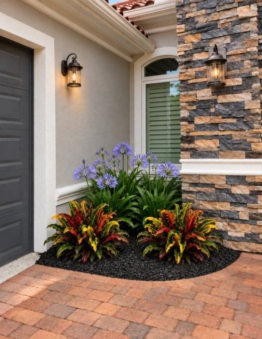 Front entry with paver walkway, dark door, wall lights, and colorful flowers beside stone facade.