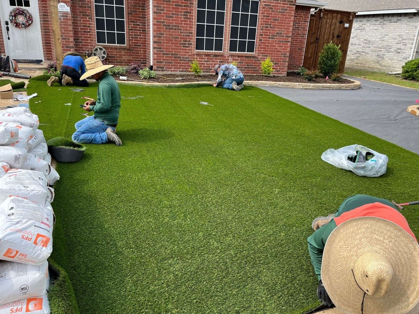 Workers installing green artificial turf in a backyard beside a brick house