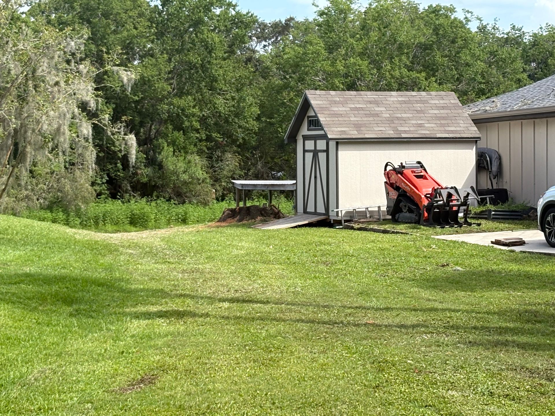 Grassy yard beside a shed with a red tractor and a parked car, backed by trees and a house.