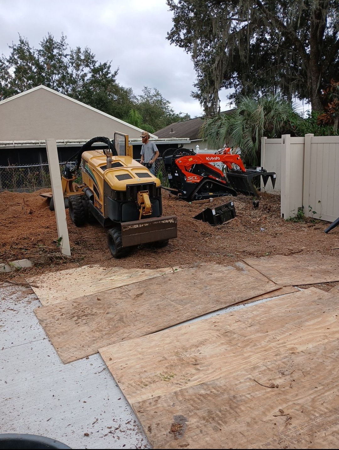 Yellow and orange skid steers parked on a gravel driveway beside a house and fence