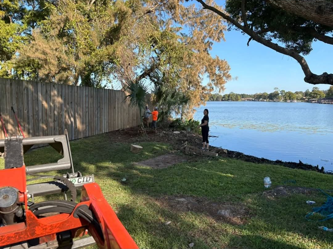 Quiet lakeside yard with a wooden fence, tree shade, and two people near the water beside an orange tractor.