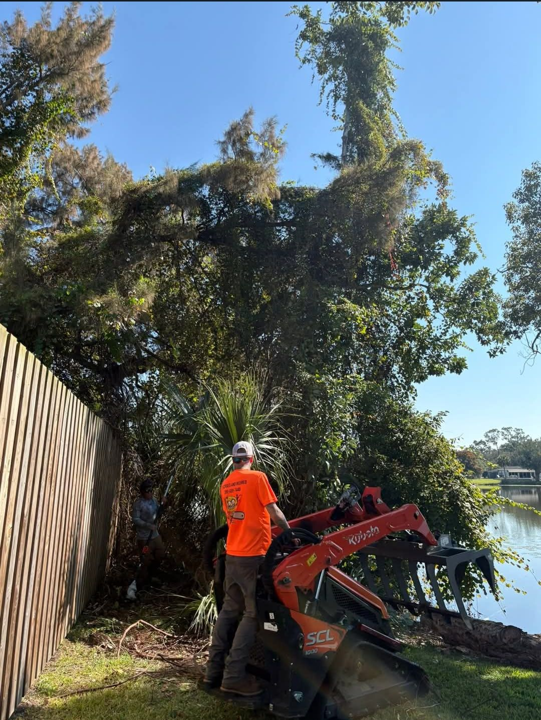 Worker in orange shirt operating a red machine near a fence beside trees and water