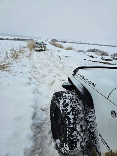 White Jeep on a snowy trail, another vehicle ahead. Winter landscape with snow-covered ground and dry grass.