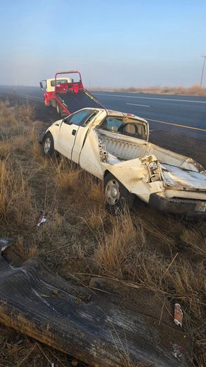Wrecked white pickup truck on side of road being towed by a red and white tow truck.