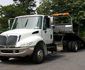 White tow truck parked on a road with an open ramp, trees in the background, and an orange light bar on top.