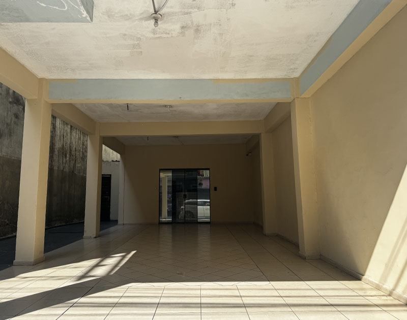 Uma ampla entrada revestida com azulejos que leva a uma porta de vidro sob um teto de concreto sustentado por pilares bege.