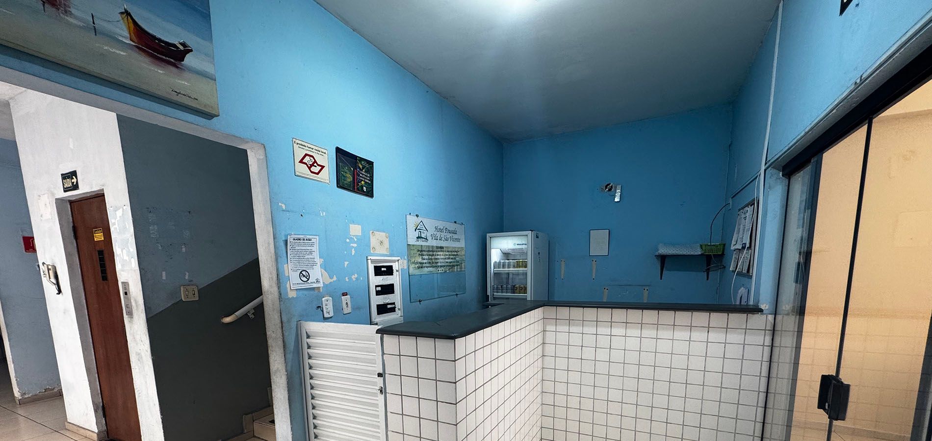 Vista interna de uma pequena área de serviço com paredes azul-claras, um balcão de recepção com azulejos brancos e uma porta à esquerda.
