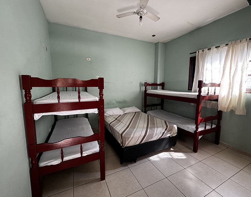 Um quarto com paredes verde-claras, um beliche à esquerda, uma cama de casal no centro e um beliche junto à janela.