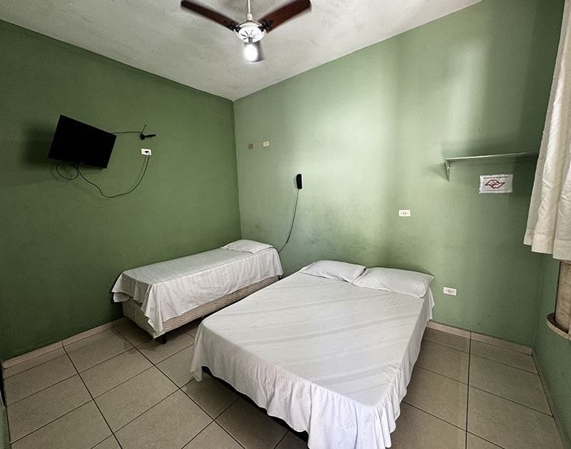Um quarto de hotel com paredes verde-sálvia, contendo uma cama de casal e uma cama de solteiro, ambas com lençóis brancos lisos.