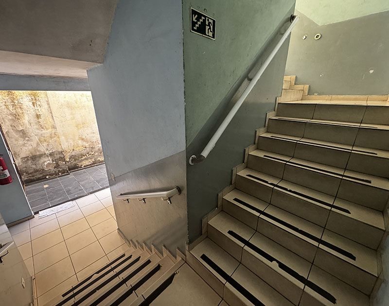 Uma escadaria com degraus revestidos de azulejos bege, com faixas antiderrapantes pretas e corrimãos de metal, que leva a uma porta de entrada ao nível da rua.