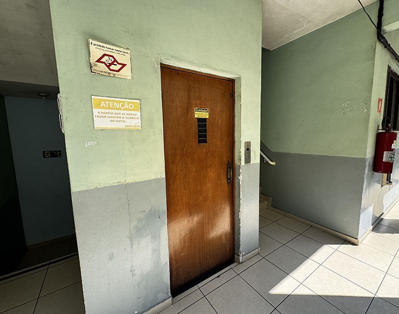 Uma porta de elevador de madeira em um corredor com paredes verde-claras e cinzas, com sinalização e um extintor de incêndio vermelho nas proximidades.