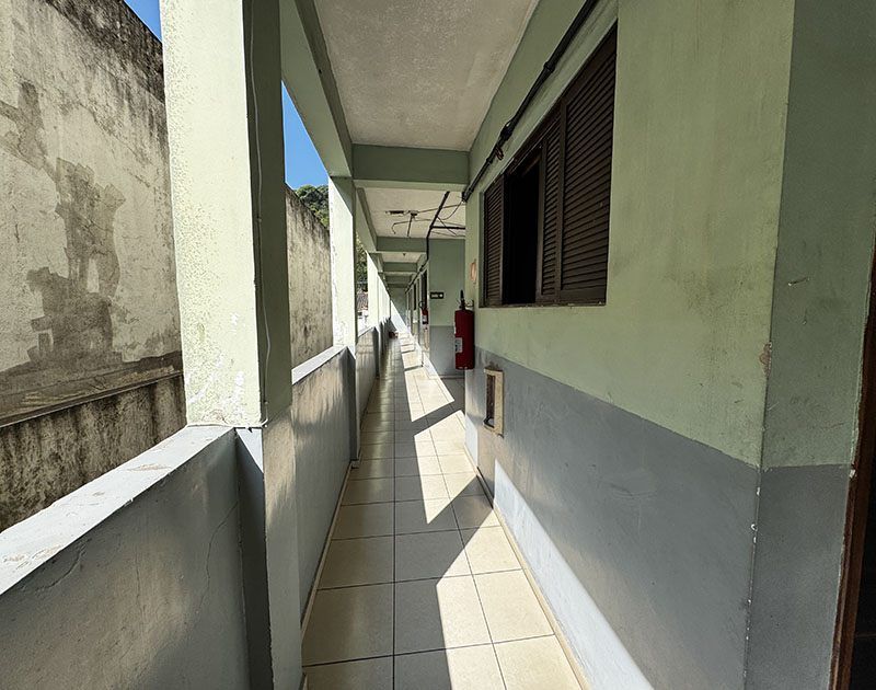 Um corredor externo com piso de azulejos, pilares de concreto e paredes em tons de verde claro e cinza, sob um céu azul límpido.