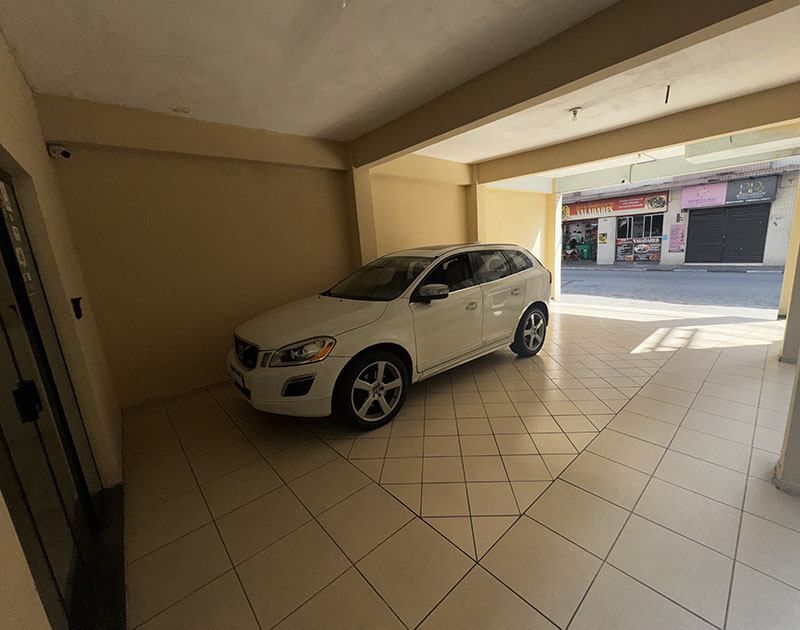 Um SUV branco estacionado em uma garagem coberta com piso de azulejo, com vista para a rua ao fundo.