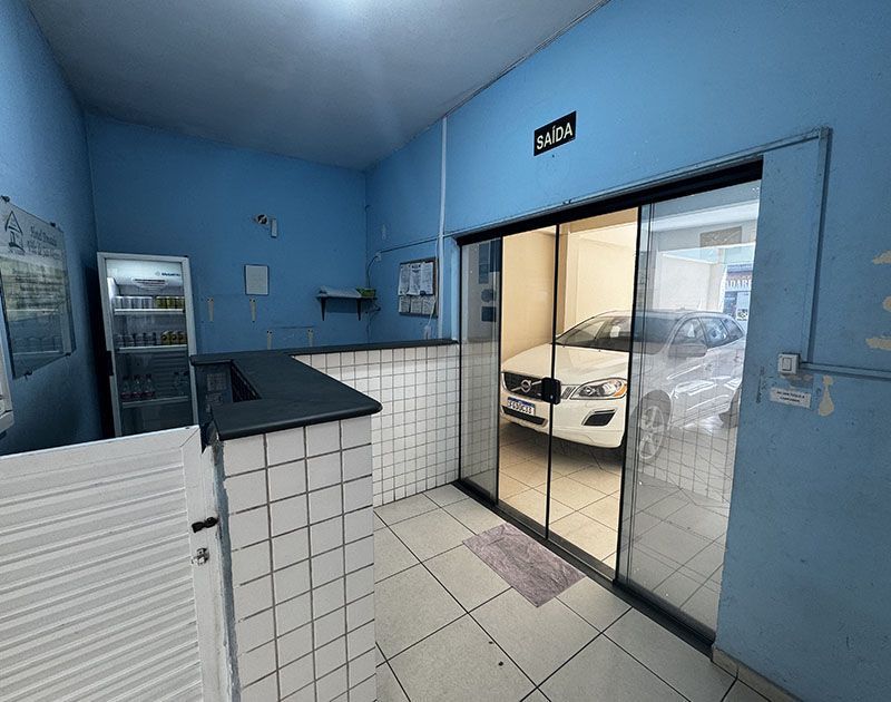 Uma sala azul-clara com um balcão de atendimento revestido de azulejos, um refrigerador expositor e uma porta de vidro deslizante que revela um carro branco.
